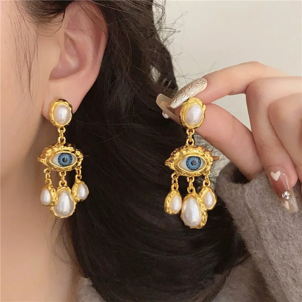 Gold and blue eye pearl drop earrings with dangling charms worn by a person's ear.