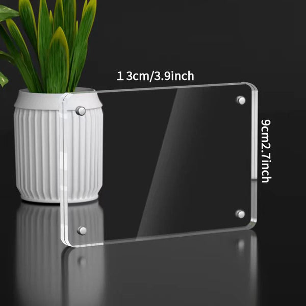A transparent acrylic photo frame with a magnetic display stand and a white cylindrical pot of green plants.