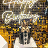 A cake decorated with a bow and presented in front of a gold sequin wall backdrop with a glowing birthday sign.