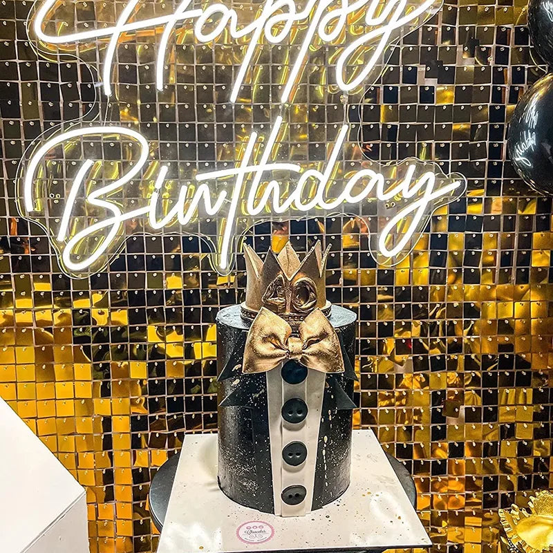 A cake decorated with a bow and presented in front of a gold sequin wall backdrop with a glowing birthday sign.