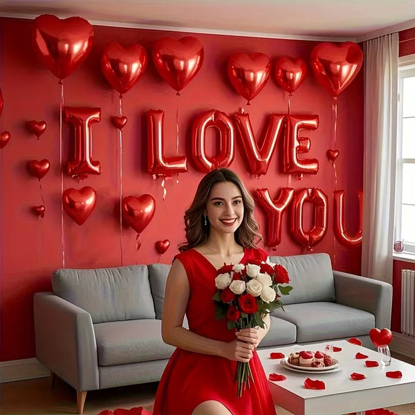 A woman in a red dress stands in front of a wall decorated with red heart foil balloons spelling out I LOVE YOU