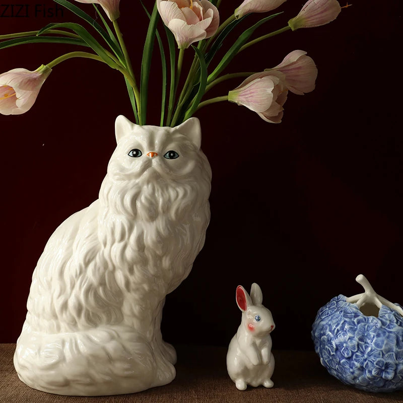 A white ceramic cat figurine sits next to a vase holding pink tulips on a dark background.