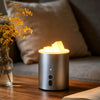 A silver electric candle warmer lamp with a warm glow illuminates several wax melts on a wooden table.