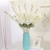 Un vase rempli de fleurs artificielles de delphinium blanches sur une surface blanche à côté d'un panier en fil de fer.