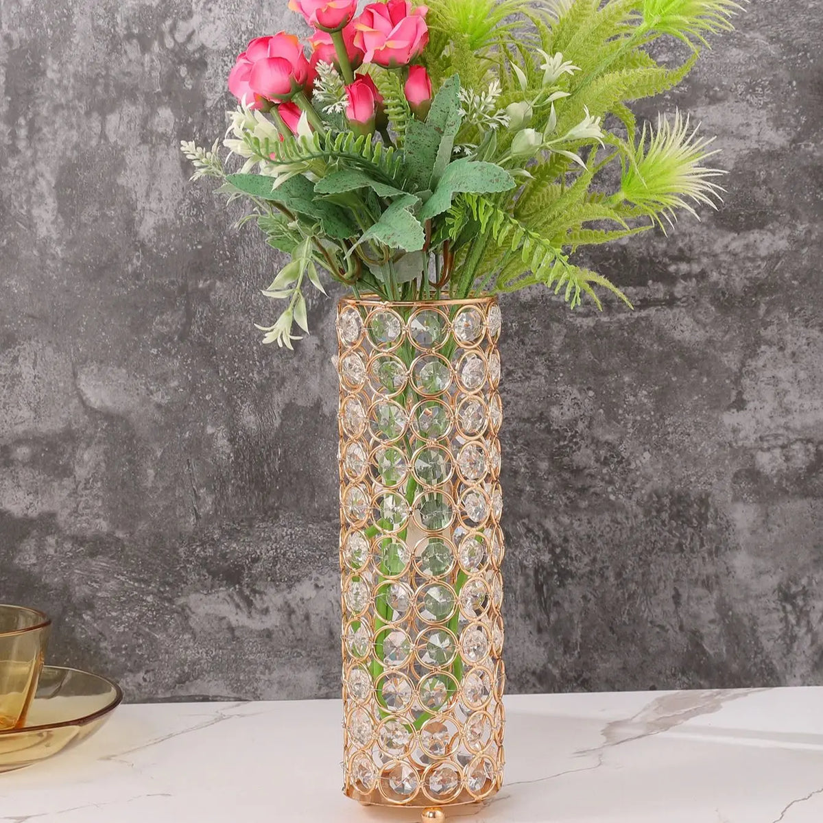 A tall crystal gold flower vase filled with pink roses and green foliage