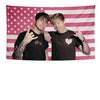 Two young men pose in front of an American flag tapestry with stars and stripes.