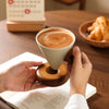 A hand holds a ceramic coffee mug with a wooden base, showing latte art on the foam.