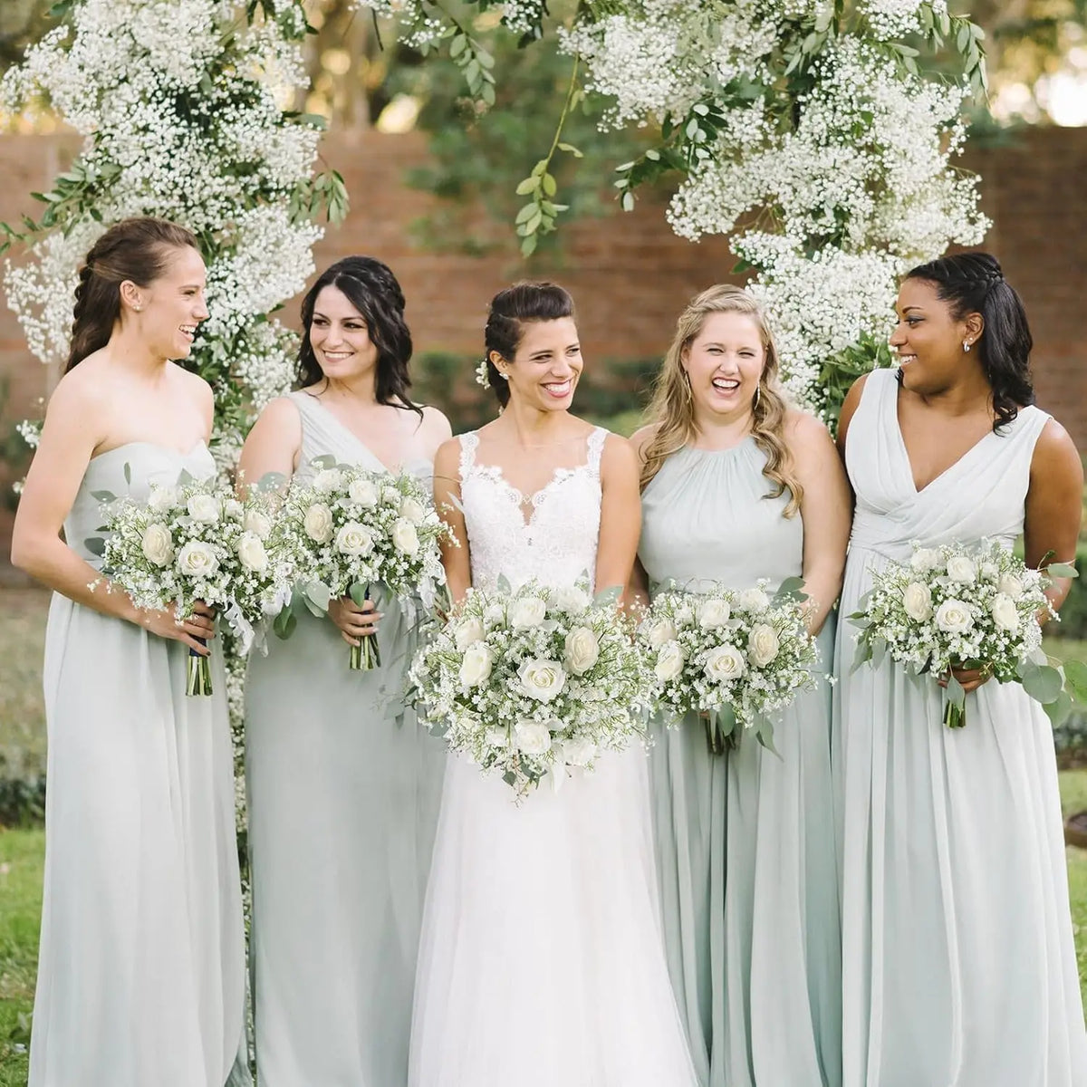 A group of women in light green dresses posing outdoors surrounded by white baby's breath flowers.