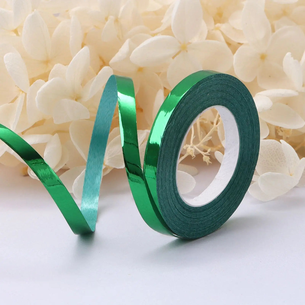 A roll of green satin balloon ribbon lying on a white surface next to white flowers.