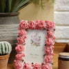 A pink rose resin picture frame surrounded by artificial roses, resting on a wooden surface.