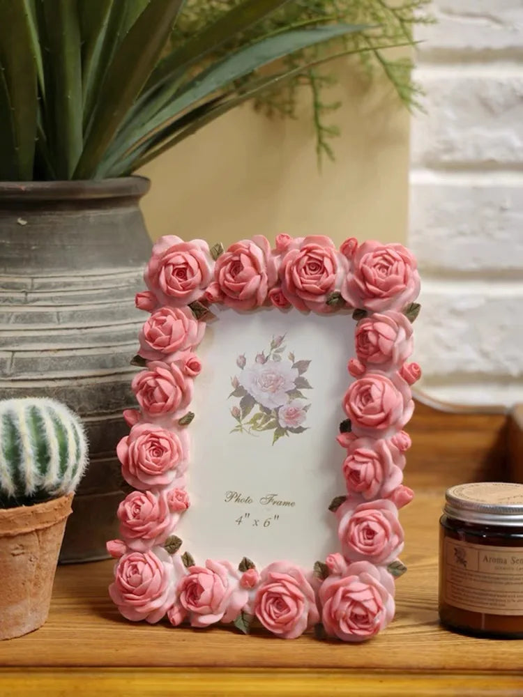 A pink rose resin picture frame surrounded by artificial roses, resting on a wooden surface.
