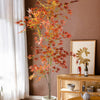 A tall artificial maple tree with reddish-orange silk leaves displayed in a terracotta pot against a brown wall.