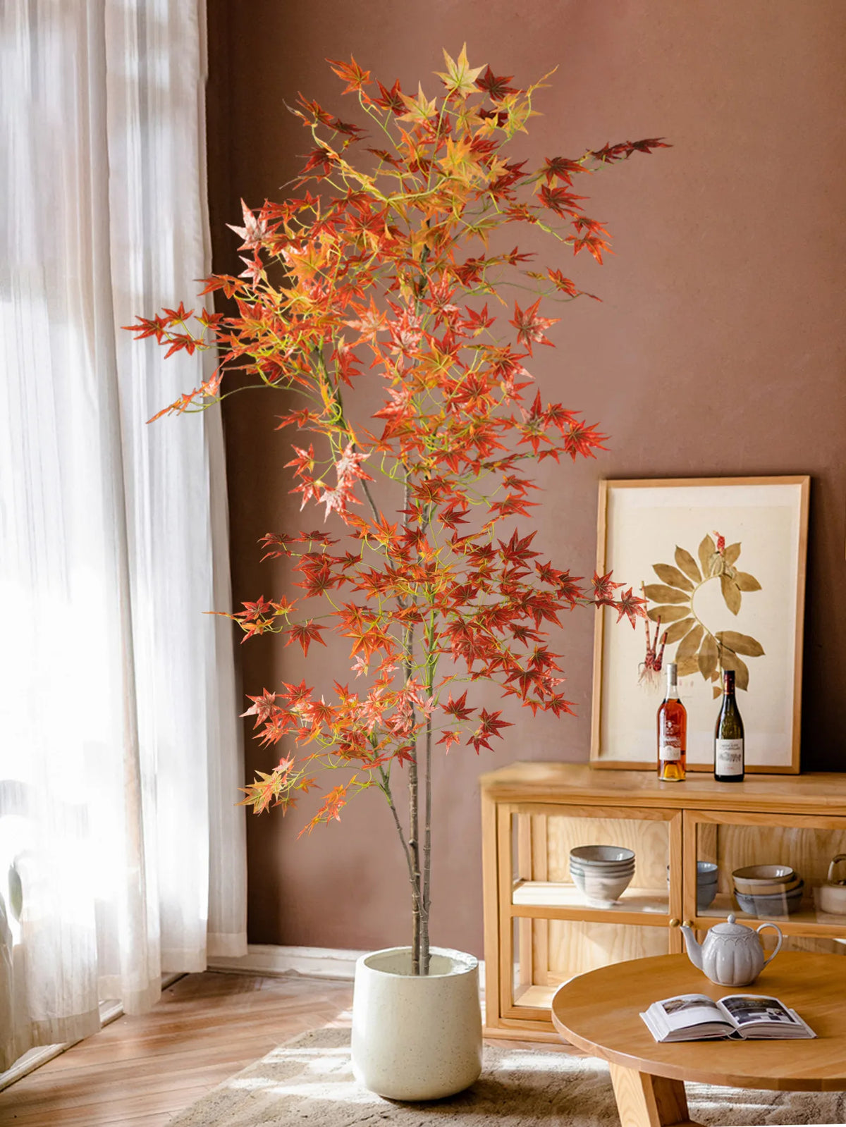 A tall artificial maple tree with reddish-orange silk leaves displayed in a terracotta pot against a brown wall.