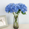 A bouquet of artificial blue hydrangea flowers in a clear glass vase on a white surface.