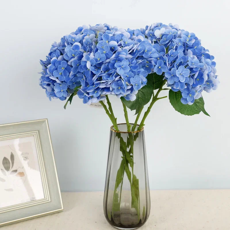 A bouquet of artificial blue hydrangea flowers in a clear glass vase on a white surface.