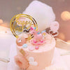 A pink cake decorated with edible flowers and a unicorn figurine, surrounded by a large white cloud and gold accents.