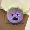 A purple plush toy resembling a monster face with green spikes is resting on brown paper against a light yellow plate.