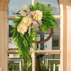 A rustic wreath of green foliage and cream hydrangeas tied with a burlap bow displayed in a window frame.
