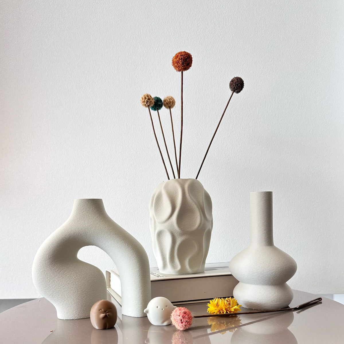 A white ceramic vase set featuring three dried floral spheres in varying shades of orange and brown on a light surface.