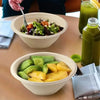 A bowl of sliced avocado and yellow fruit served on a table with a glass of green juice.