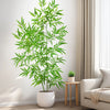 An artificial bamboo branch with green leaves displayed in a white pot on a wooden stand next to a light-colored sofa.