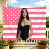 A woman wearing a black strapless dress stands in front of a pink and white striped flag tapestry.