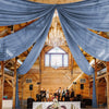 Blue sheer chiffon curtains drape across the rafters of a wooden barn interior for an event.
