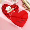 A red heart-shaped gift box filled with red roses and a white plush bear, tied with a red ribbon.
