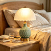 A ceramic table lamp with a pale green crackle glaze sits on a wooden nightstand next to a bed.