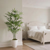 A tall artificial bamboo tree in a white pot placed next to a light wood bed with white linens.