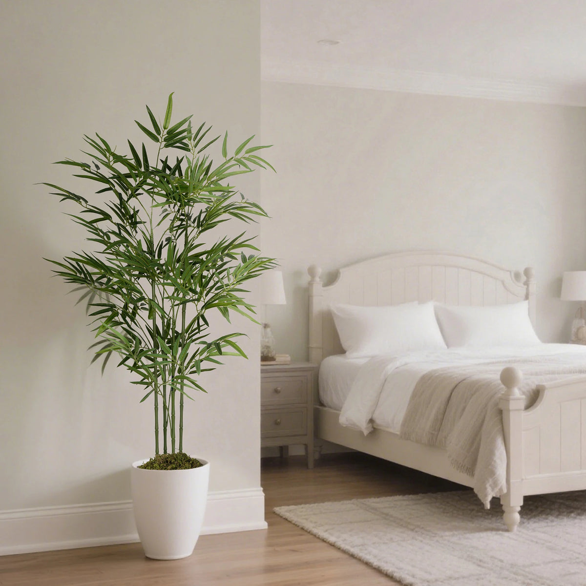 A tall artificial bamboo tree in a white pot placed next to a light wood bed with white linens.