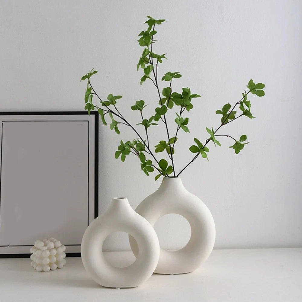 A white ceramic vase holds a sprig of green foliage against a white wall.