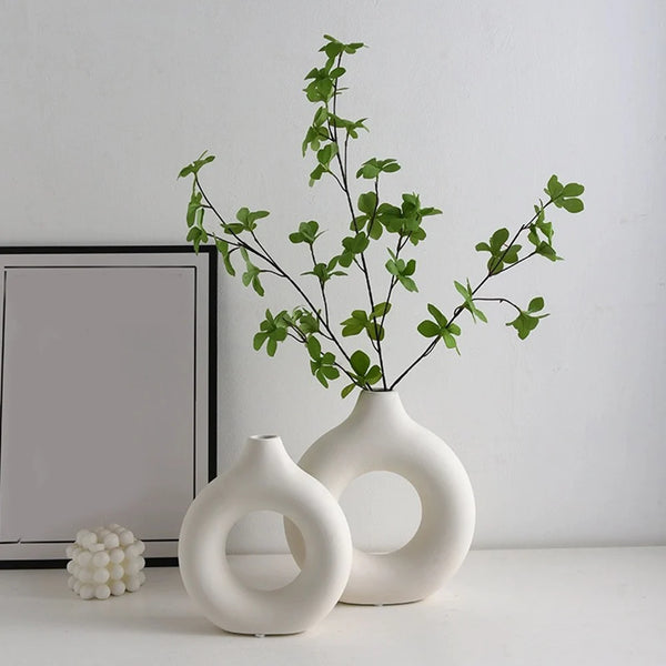 A white ceramic vase holds a sprig of green foliage against a white wall.