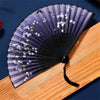 A purple and black vintage silk folding hand fan with white floral designs resting on a wooden surface.