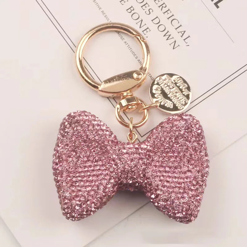A sparkling pink rhinestone bow keychain with a gold-toned clasp and attached to a white card.
