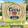 A beige flag banner featuring the text Coors Banquet since 1873 and 150 years, set outdoors on a grassy lawn.