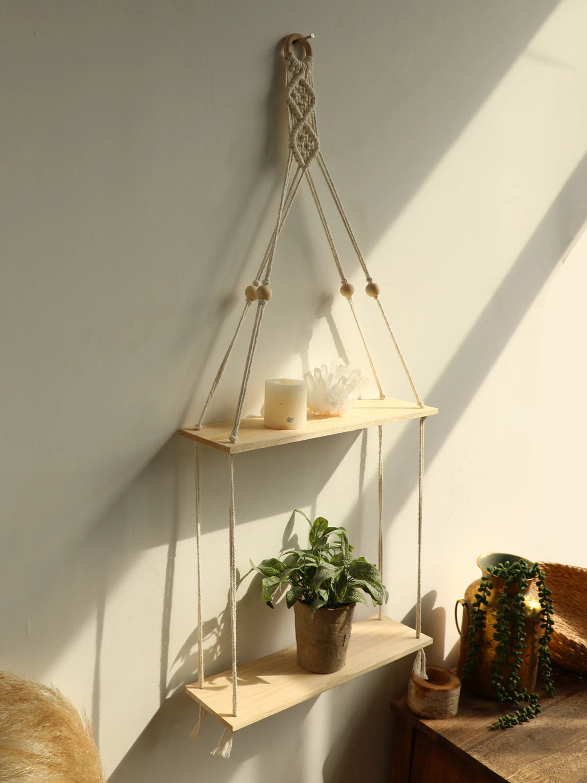 A macrame wood floating wall shelf with hanging rope displaying a candle and a potted plant on a light-colored wall.