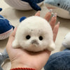 A hand holds a small white seal plush keychain against a blurred background of other stuffed animals.