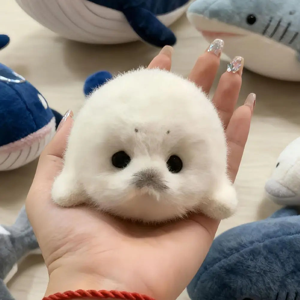 A hand holds a small white seal plush keychain against a blurred background of other stuffed animals.