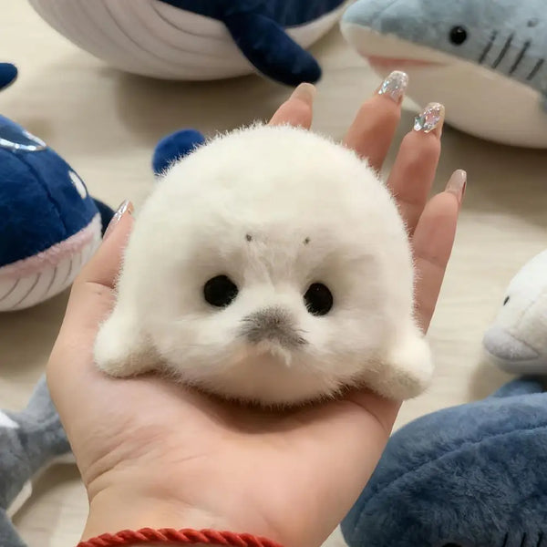 Une main tient un petit porte-clés en peluche de sceau blanc contre un arrière-plan flou d'autres animaux en peluche.