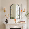 An arched wall mirror with an aluminum frame hangs above a marble mantelpiece.