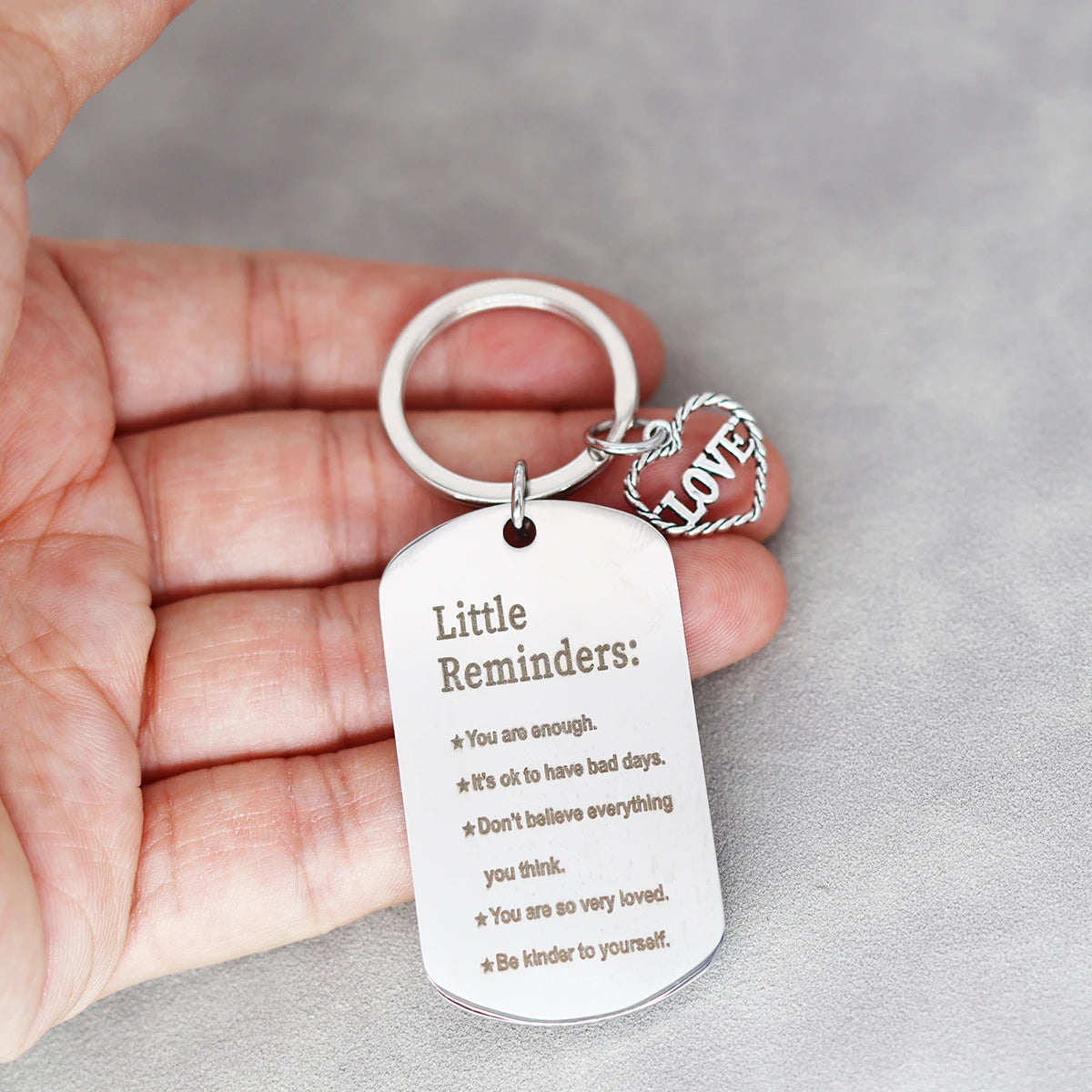 A stainless steel heart-shaped keychain with inspirational reminders engraved on it, held in a hand.