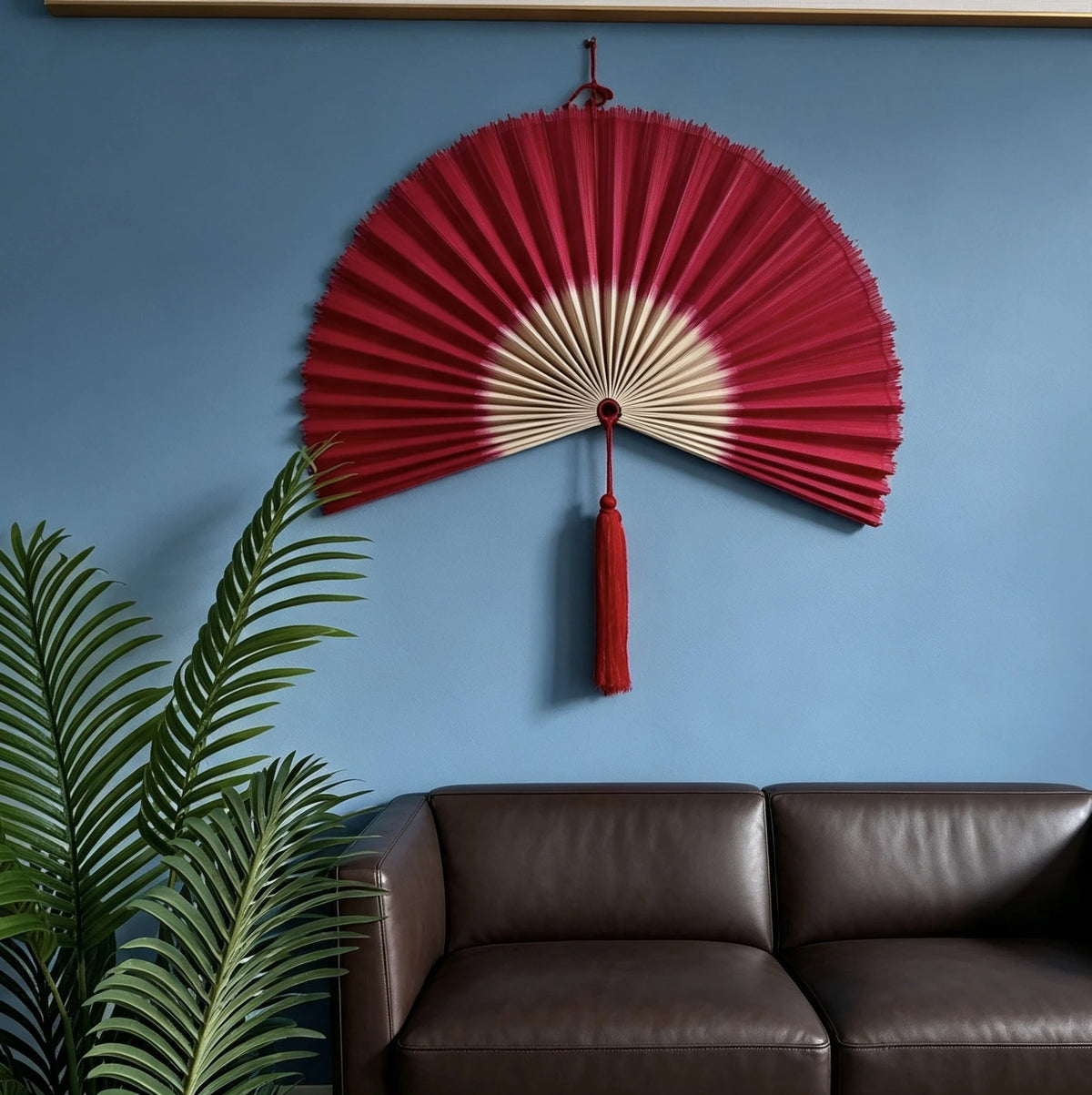 A large red and cream handwoven bamboo wall fan decor with a tassel mounted on a blue wall above a brown leather sofa and gre