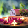 Pink floating candles on a wooden plate surrounded by dark petals and green leaves