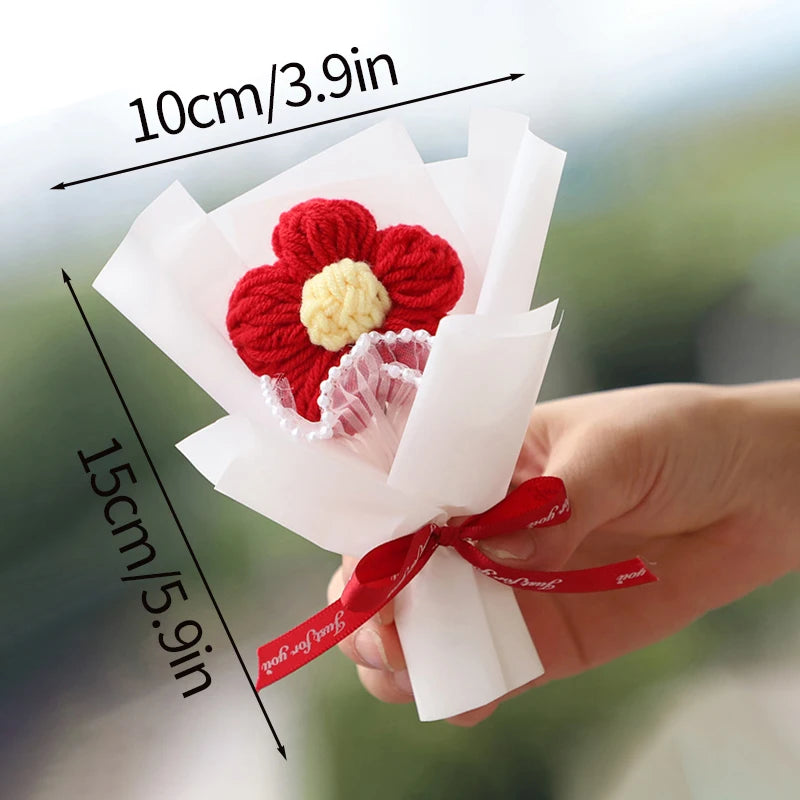 A hand holds a bouquet of handmade red and yellow crochet flowers wrapped in white paper.