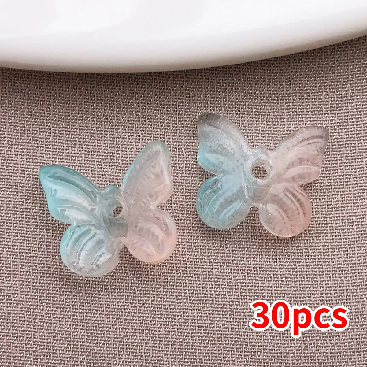 Colección de cuentas de mariposa de vidrio azul claro y rosa translúcido esparcidas sobre una superficie gris texturizada.