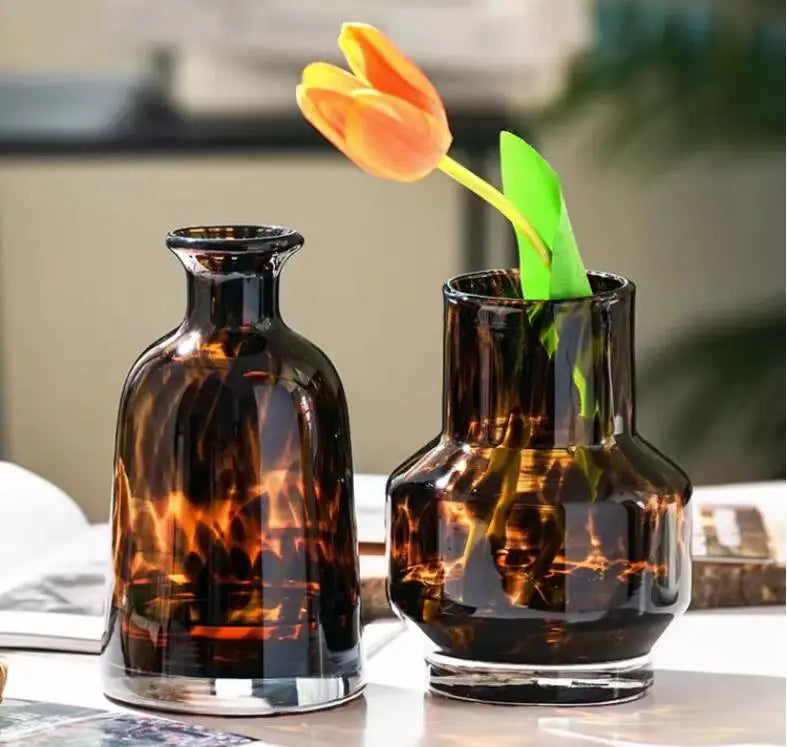 A dark amber glass vase holds a single orange tulip with a green stem.