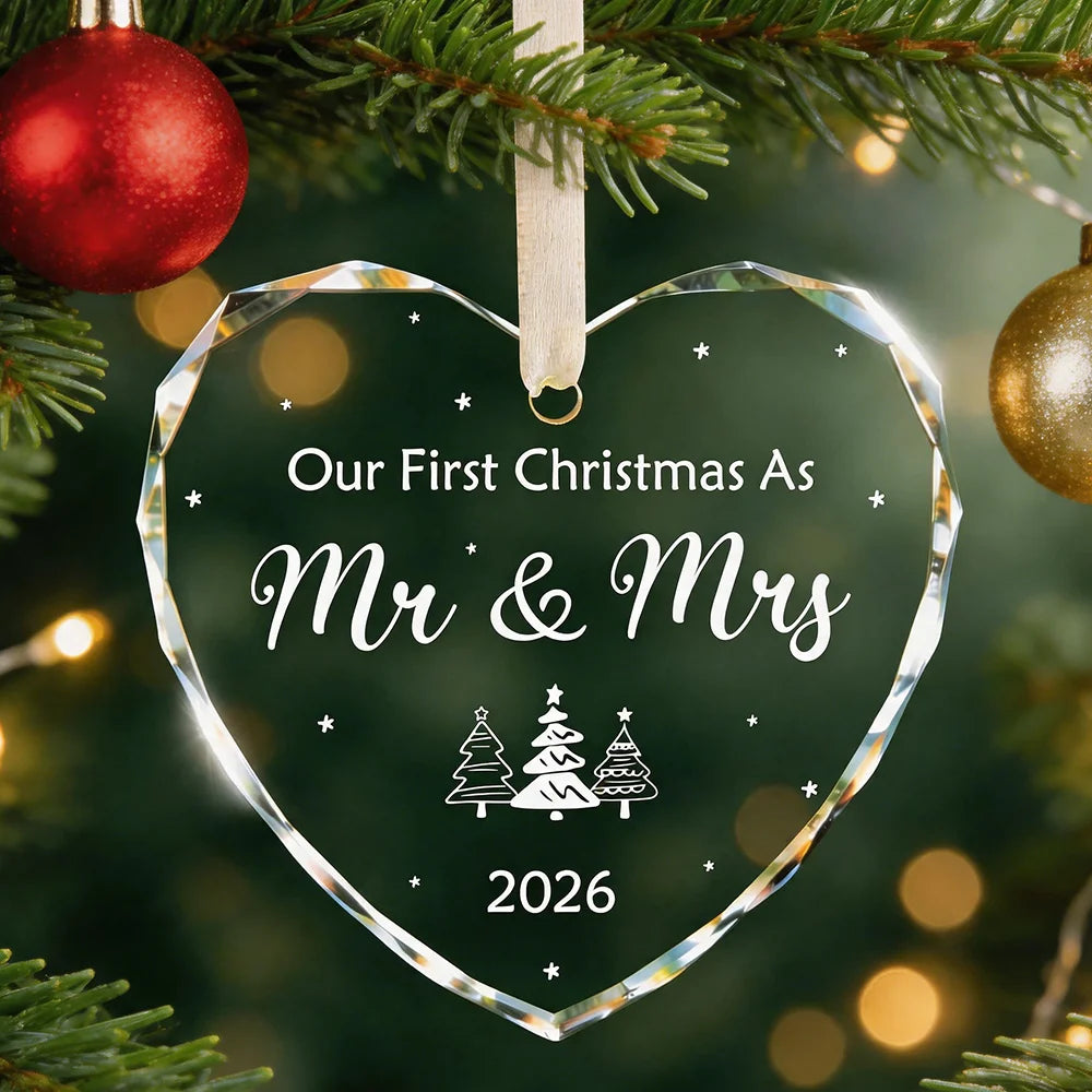A clear heart-shaped crystal ornament with the text Our First Christmas As Mr & Mrs 2026 hangs on a green Christmas tree bran