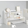 A white two-layer desktop organizer shelf displaying various small items like bottles, candles, and accessories.