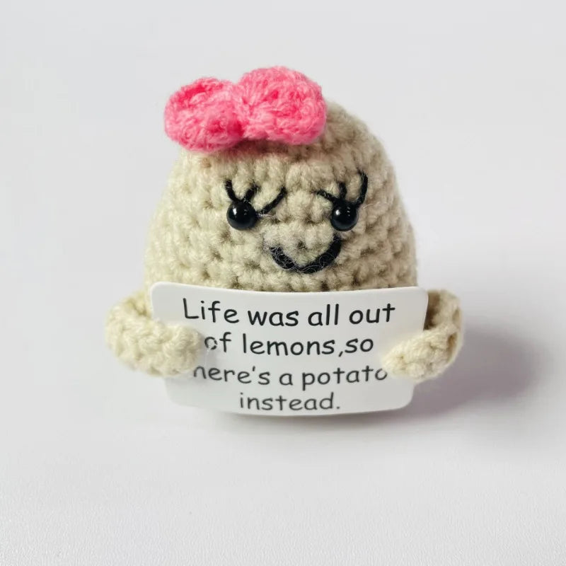 A handmade knitted plush doll with light beige skin and a pink bow, holding a white sign that reads Life was all out of lemon